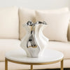 A white ceramic vase with a wave pattern sits on a round gold table in a bright living room setting.