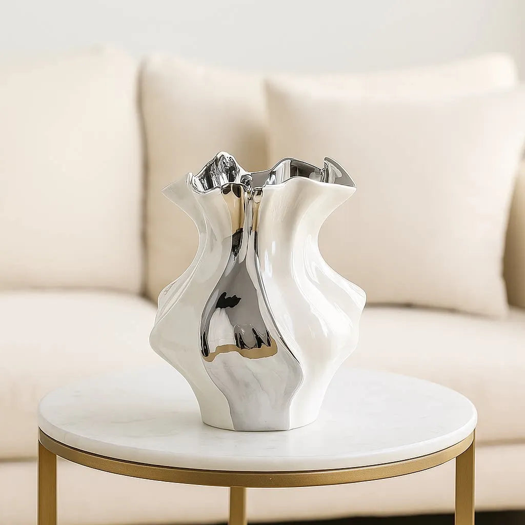 A white ceramic vase with a wave pattern sits on a round gold table in a bright living room setting.