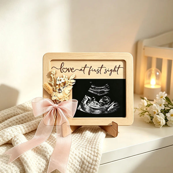 A wooden ultrasound photo frame with a black display and a pink ribbon tied around it, resting on a white surface.