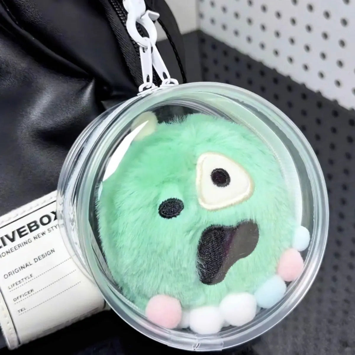 A light green plush doll keychain charm is displayed in a clear plastic container against a dark background.