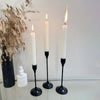 A set of three white taper candles in black holders displayed on a glass surface next to dried foliage.