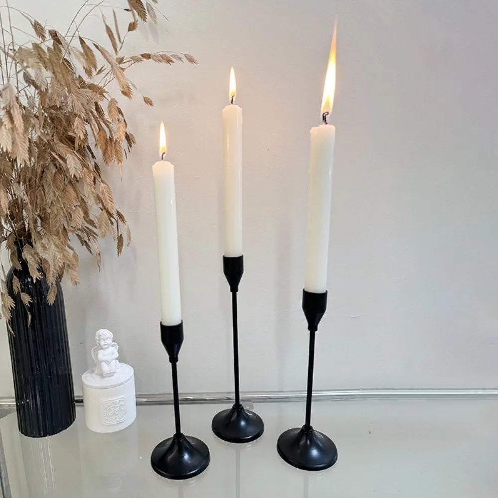 A set of three white taper candles in black holders displayed on a glass surface next to dried foliage.