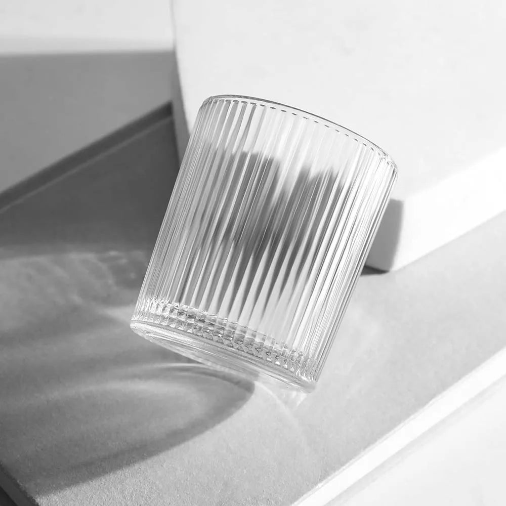 A close-up, black and white photo of a clear glass votive candle holder with vertical ridges, resting on a light surface.
