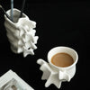 A white ceramic vase shaped like a vertebra holds a cup of coffee on a dark surface.