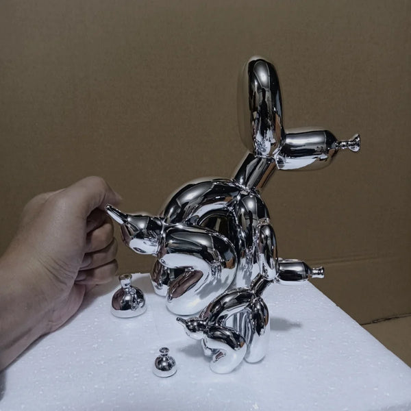 A chrome sculpture of a dog squatting, made of polished metal, resting on a white surface.