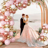 A bride and groom stand under a pink and gold balloon arch decorated with latex balloons and greenery.