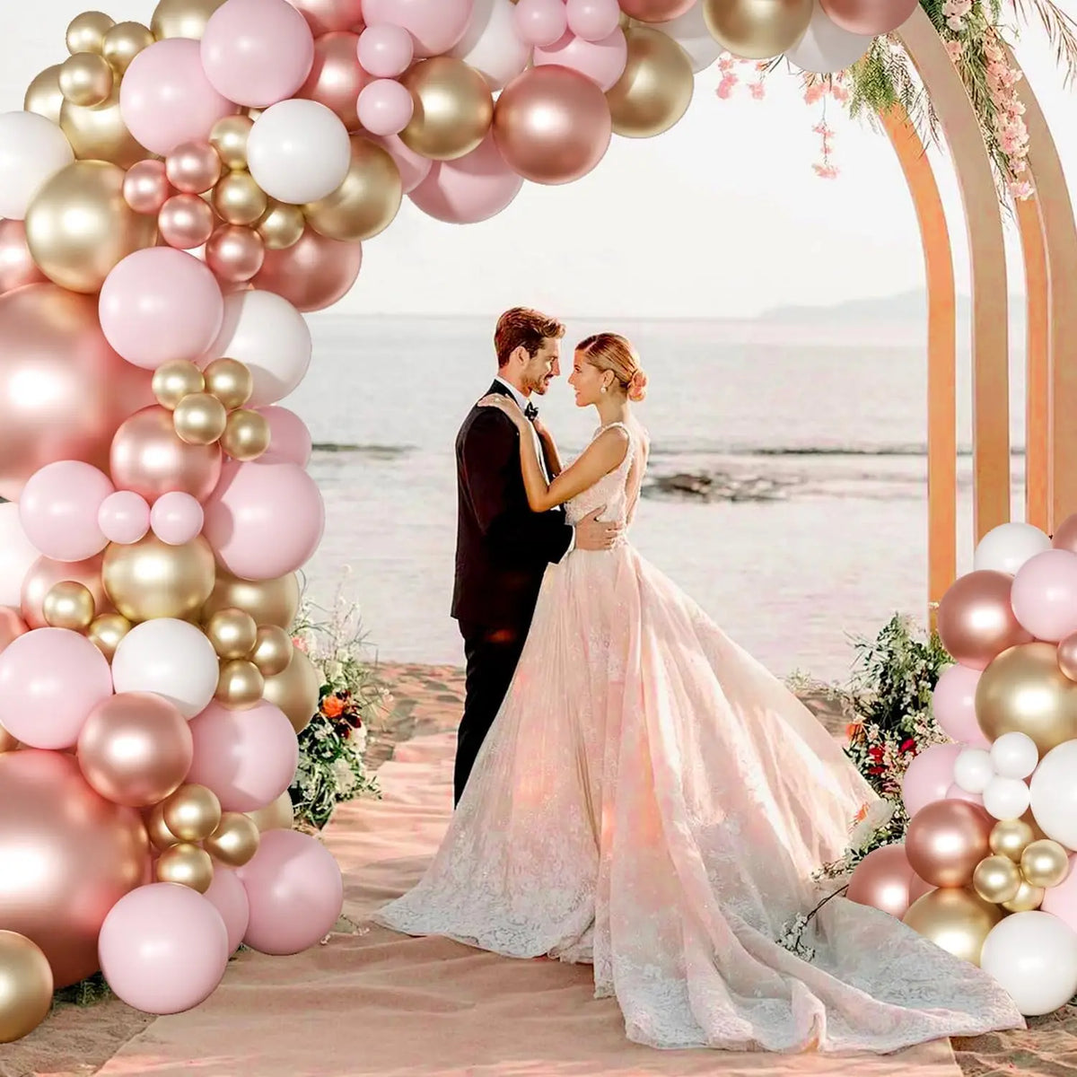 A bride and groom stand under a pink and gold balloon arch decorated with latex balloons and greenery.