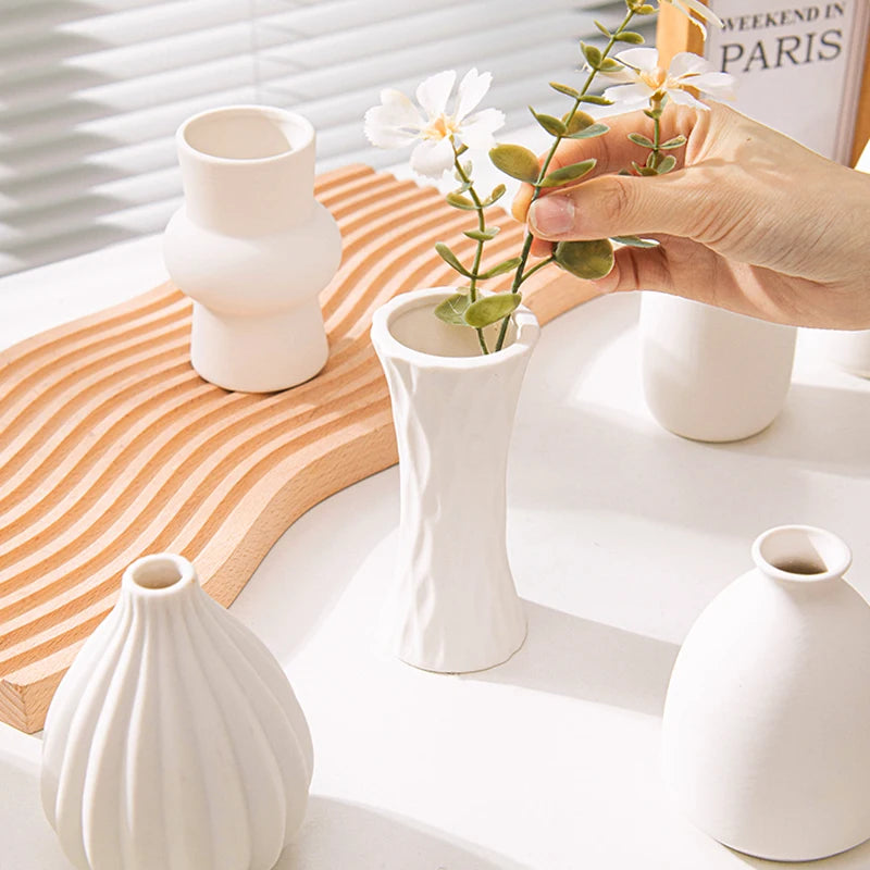 A hand holds a delicate green plant in a white ceramic bud vase on a wooden surface.