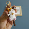 A handcrafted crochet cat keychain pendant with brown and white fur and a gold-colored ring.