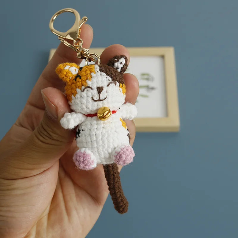A handcrafted crochet cat keychain pendant with brown and white fur and a gold-colored ring.