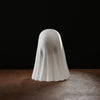 A white ghost figurine with a playful pose is displayed on a dark wooden surface against a black background.