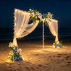 A wedding arch backdrop made of sheer fabric and string lights decorated with white and blue flowers on a beach at night