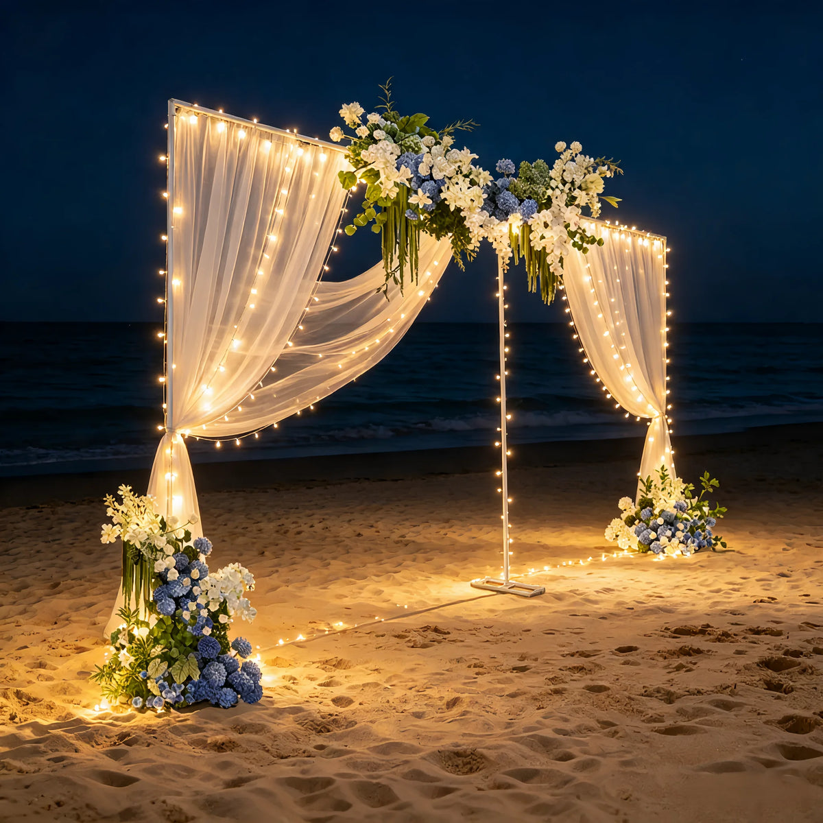 A wedding arch backdrop made of sheer fabric and string lights decorated with white and blue flowers on a beach at night