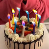 A cake decorated with vibrant flame decorative candles and fresh fruit, held by a person in a pink top.