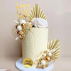 A tiered cake decorated with white, gold, and beige elements including floral accents and fan shapes.