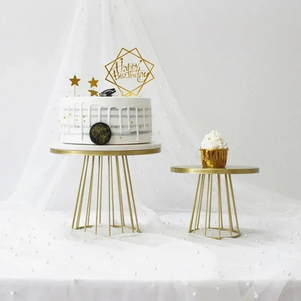 A white tiered cake with gold accents sits on a gold metal stand next to another small golden cupcake display.
