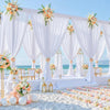 A white fabric backdrop curtain setup for an outdoor event featuring draped fabric and floral accents on a sandy beach.
