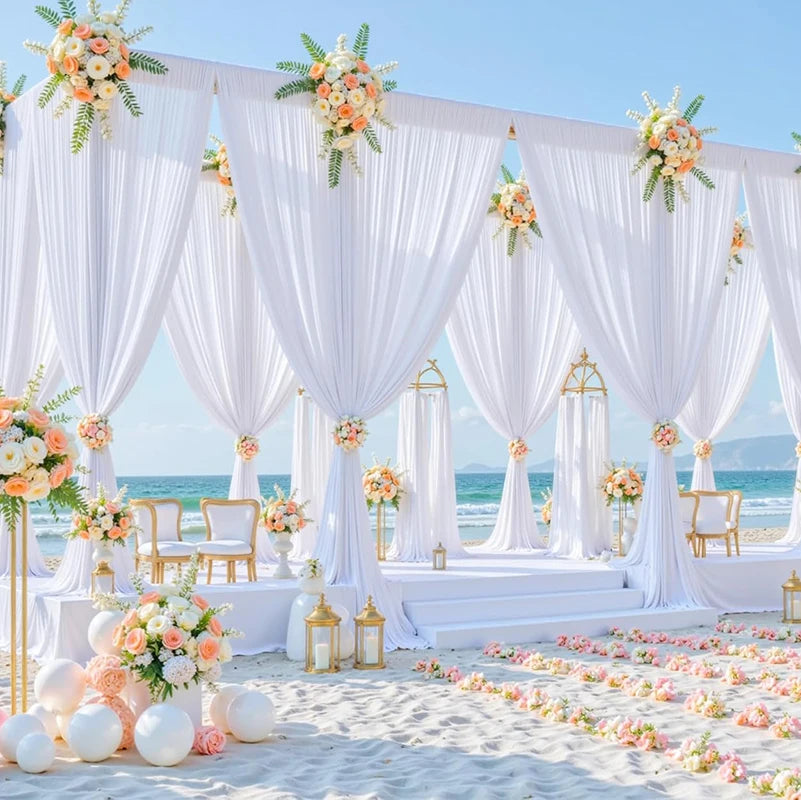 A white fabric backdrop curtain setup for an outdoor event featuring draped fabric and floral accents on a sandy beach.
