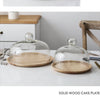 A clear glass dome cover sits on a round solid wood cake plate on a white marble surface.