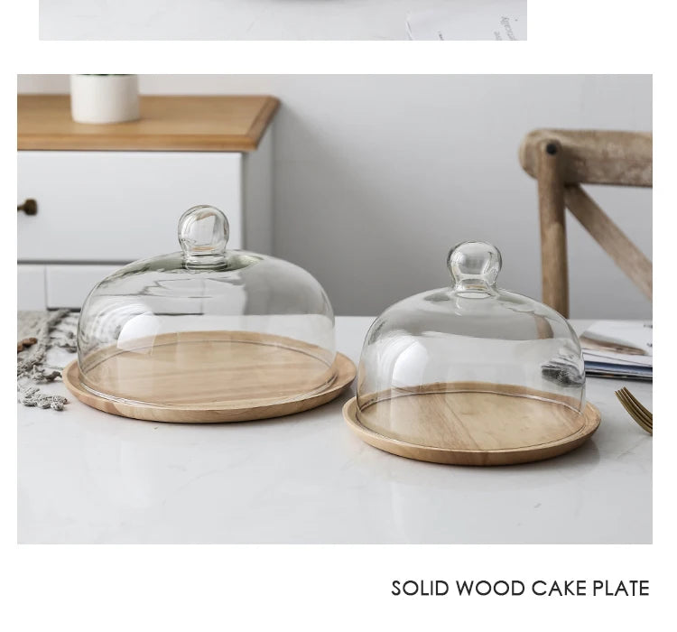 A clear glass dome cover sits on a round solid wood cake plate on a white marble surface.