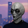 A close-up of a realistic alien full-face latex mask with large black eyes against a city skyline at sunset.