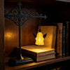 A black wrought iron cross lamp with a warm glowing glass shade sits on a wooden shelf next to stacked books.