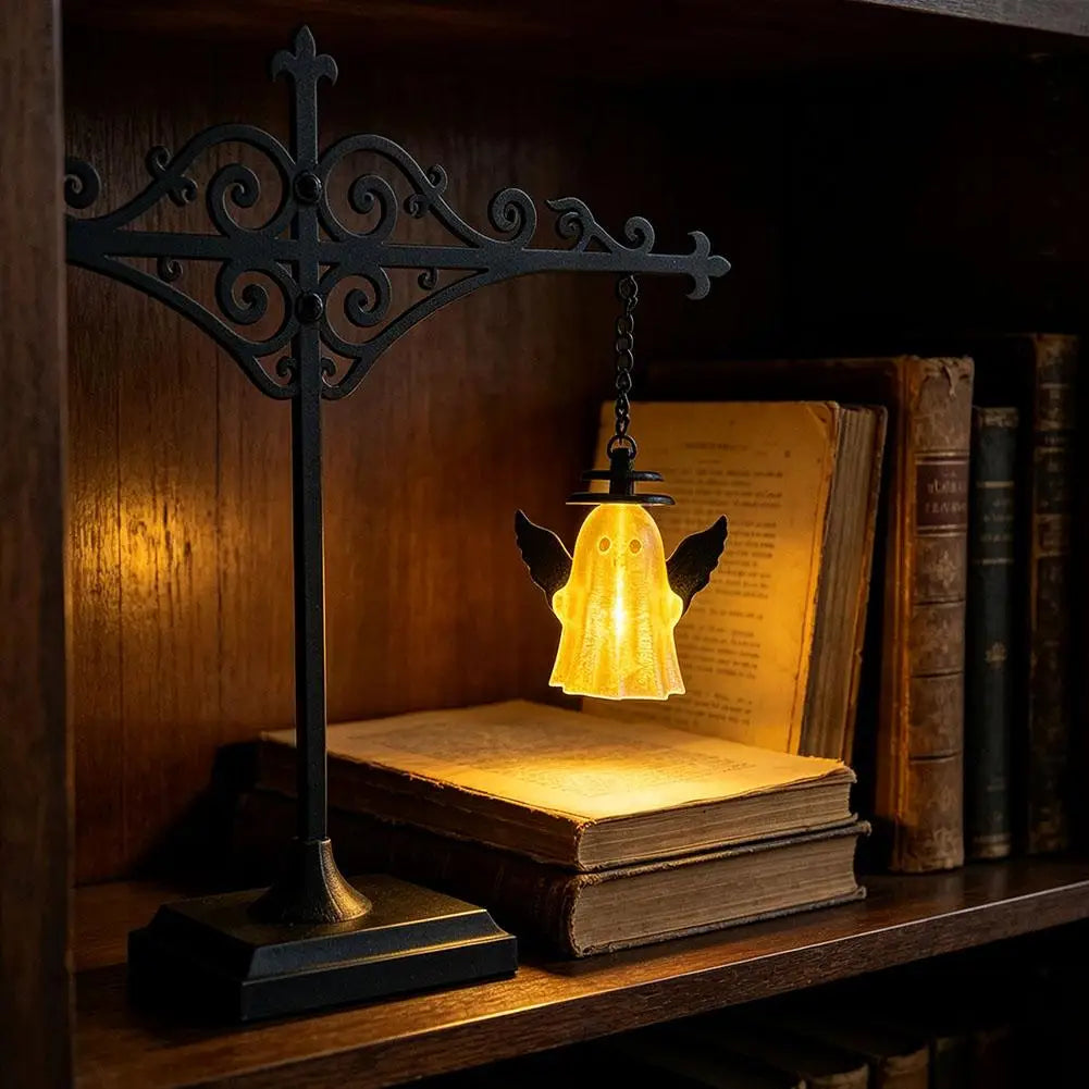 A black wrought iron cross lamp with a warm glowing glass shade sits on a wooden shelf next to stacked books.