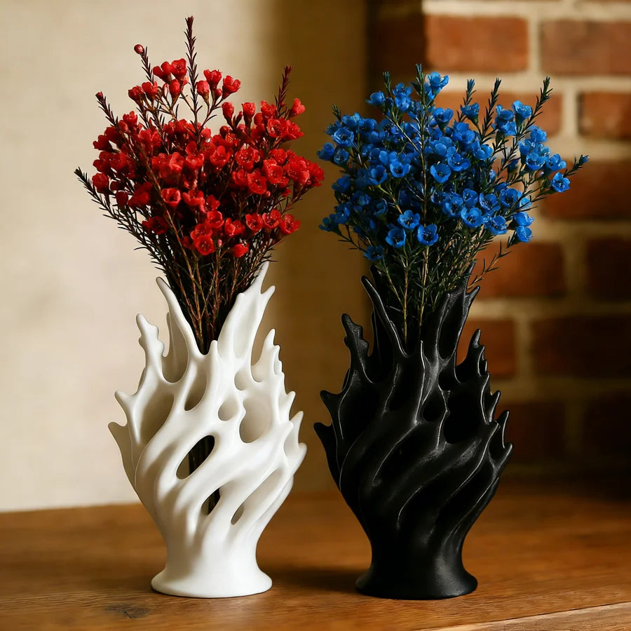 A pair of modern plastic flower vases, one white and one black, holding vibrant red and blue flowers against a brick backgrou