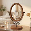 An elegant rotating oval vanity mirror with a wooden frame sits on a glass table.
