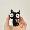 A handcrafted crochet cat keychain pendant with black fur and white eyes is held in a person's hand.