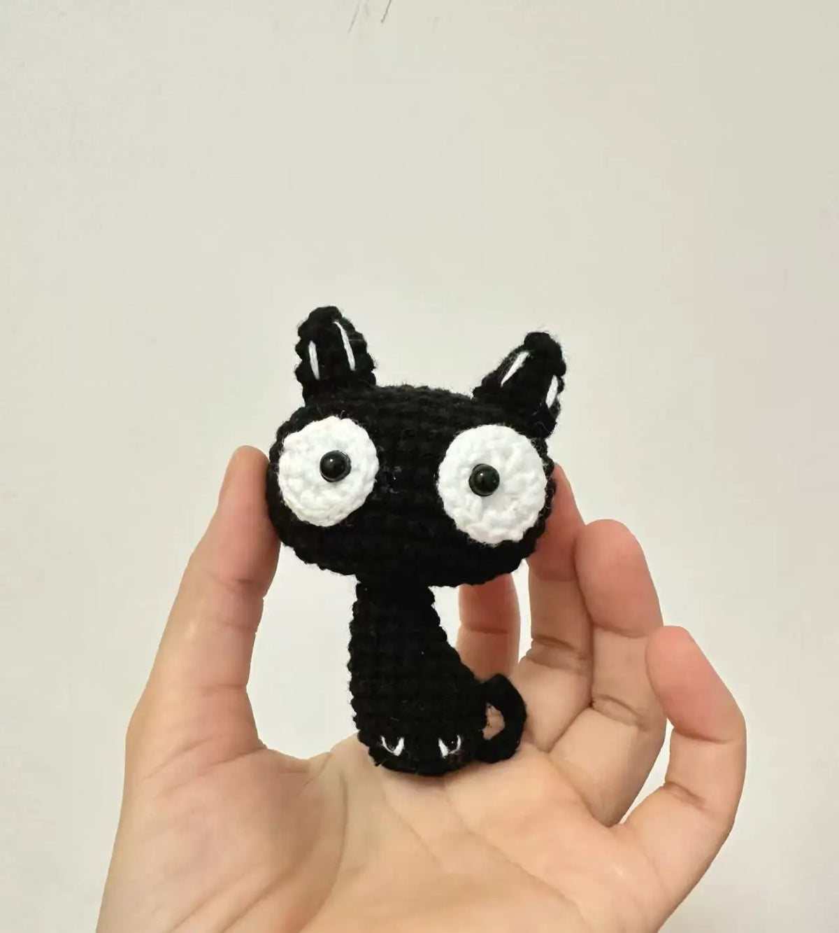 A handcrafted crochet cat keychain pendant with black fur and white eyes is held in a person's hand.