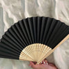 A black hand folding fan with light wood accents is held in a person's hand against a white background.