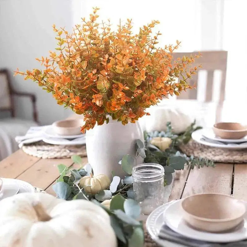 A bouquet of artificial orange and yellow fall flowers in a white vase on a wooden table.