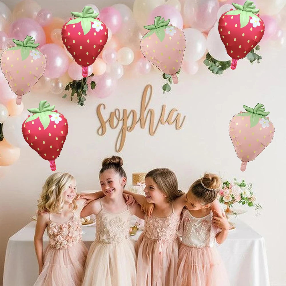 Five girls in pink dresses pose in front of a wall decorated with pink and green strawberry foil balloons and the name Sophia