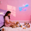 A young girl sits on the floor surrounded by stuffed animals in front of a pink wall with a neon dachshund sign.