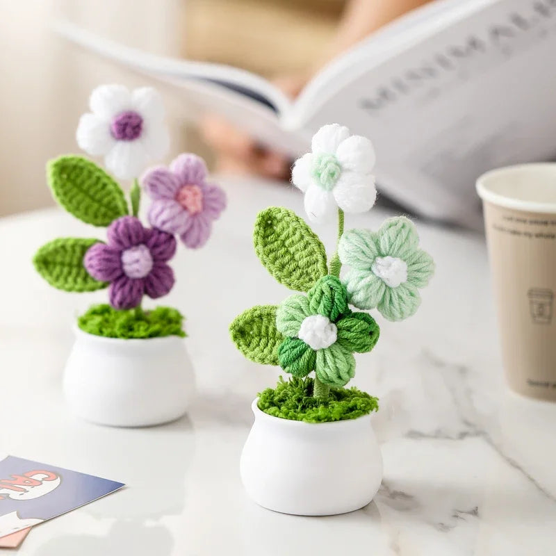 Hand woven crochet flowers in white and purple are potted in small white ceramic cups on a marble surface.