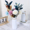 A floral arrangement featuring dark blue and muted green leaves in a white vase with a gold twine wrap.