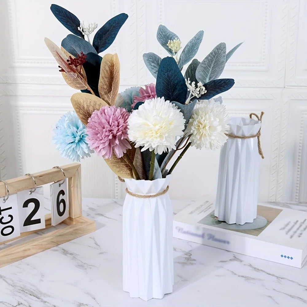 A floral arrangement featuring dark blue and muted green leaves in a white vase with a gold twine wrap.