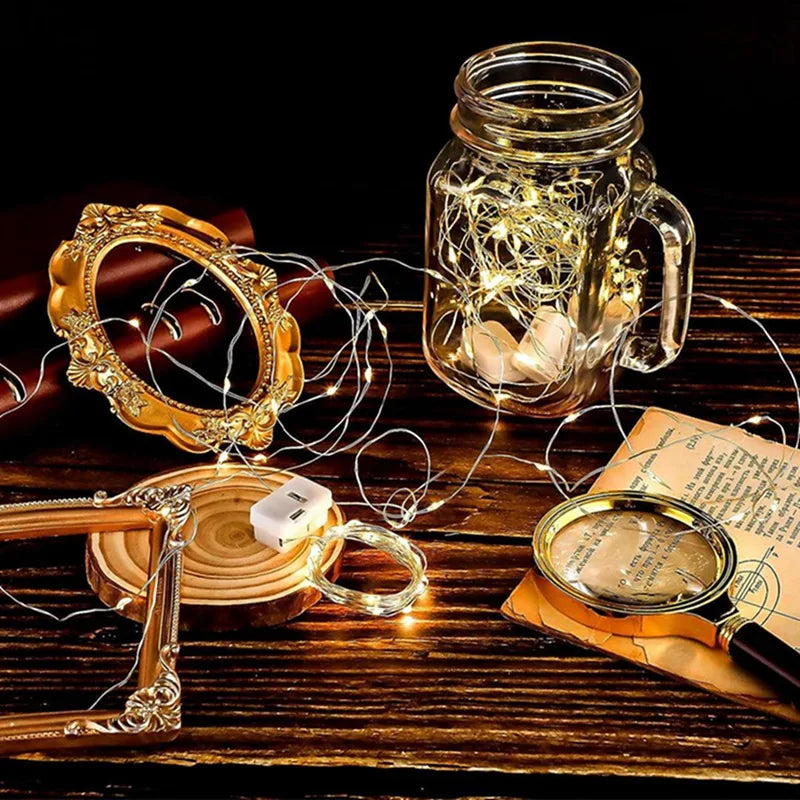 A glass jar filled with glowing copper wire fairy lights rests on a wooden surface next to a gold-colored wire wreath and a m