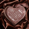A clear acrylic heart pendant engraved with 'I love you' and a rose design, resting on dark brown satin fabric.