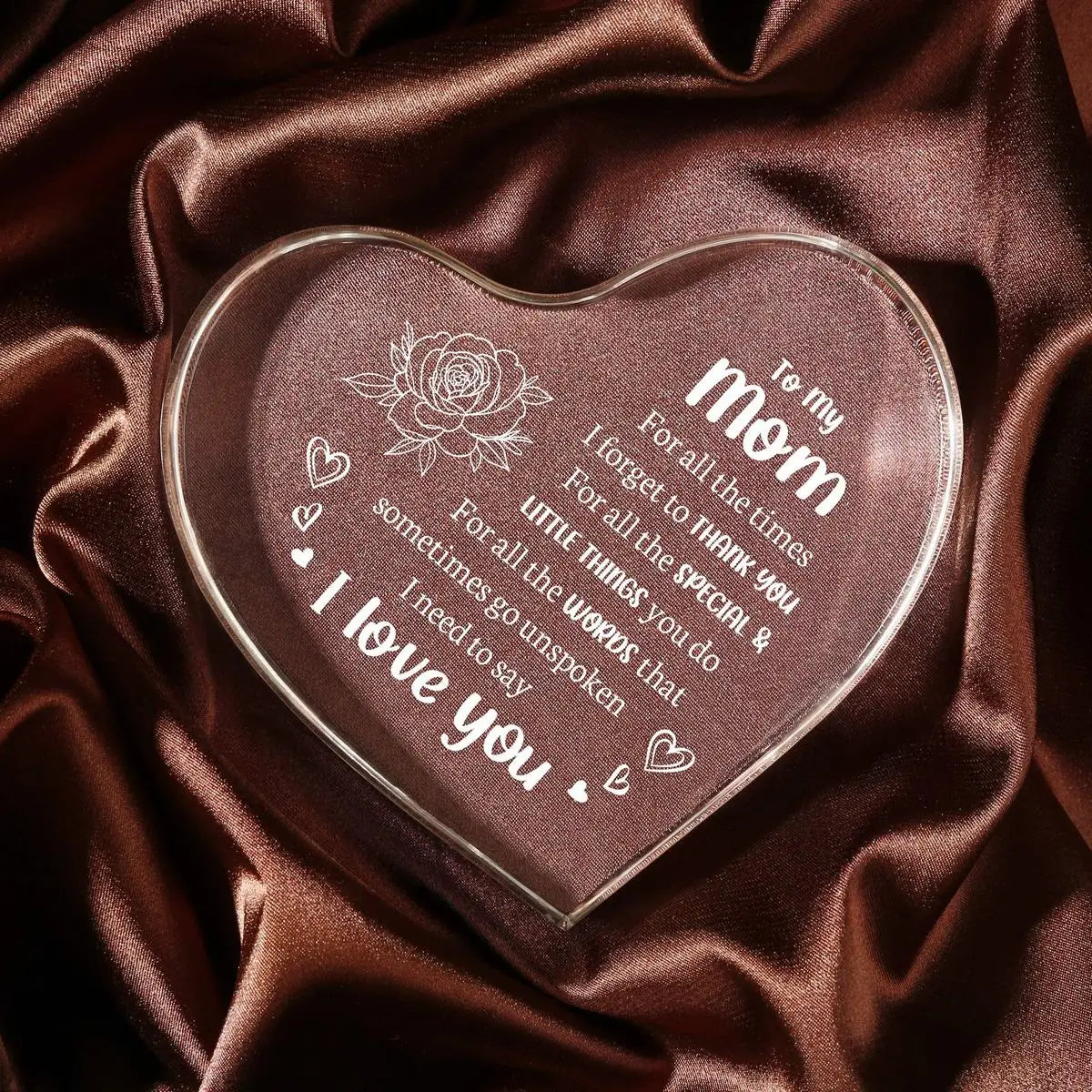 A clear acrylic heart pendant engraved with 'I love you' and a rose design, resting on dark brown satin fabric.