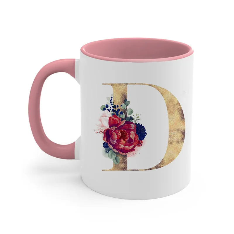 A pink ceramic coffee mug featuring a large gold letter D decorated with colorful floral accents.