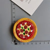 A round pizza-shaped food magnet with red tomato toppings and green olives, placed next to a ruler.