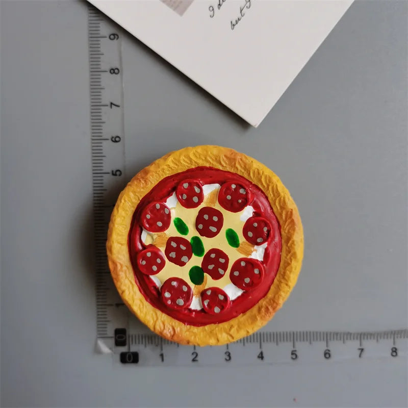 A round pizza-shaped food magnet with red tomato toppings and green olives, placed next to a ruler.