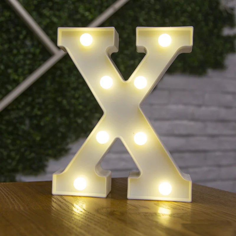 A glowing warm white LED alphabet letter light shaped like the letter X resting on a wooden surface.