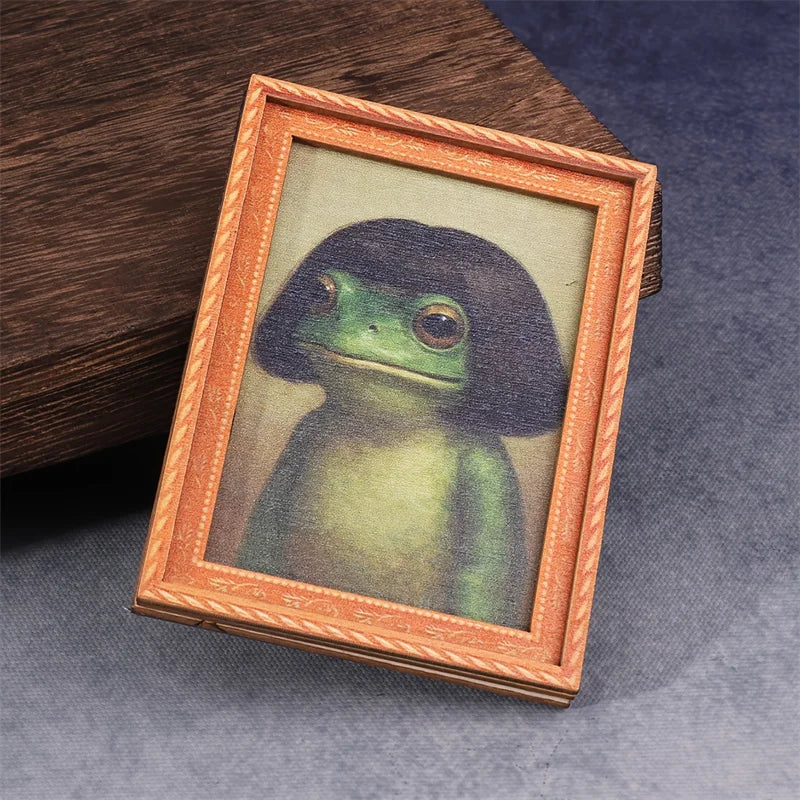 A framed illustration of a green frog in a wooden frame, displayed on a dark surface.