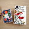 A white book-shaped ceramic vase with floral and botanical illustrations is displayed on a wooden surface.