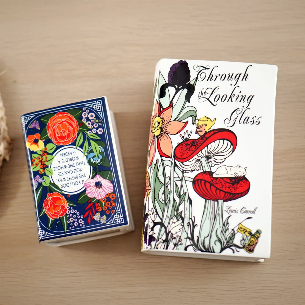 A white book-shaped ceramic vase with floral and botanical illustrations is displayed on a wooden surface.