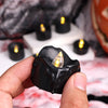 A hand holds a black wax candle with a warm white glow, set against a blurred background of other candles and dark fabric.