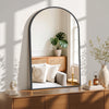 An arched wall mirror with an aluminum frame reflecting a green plant and interior light.